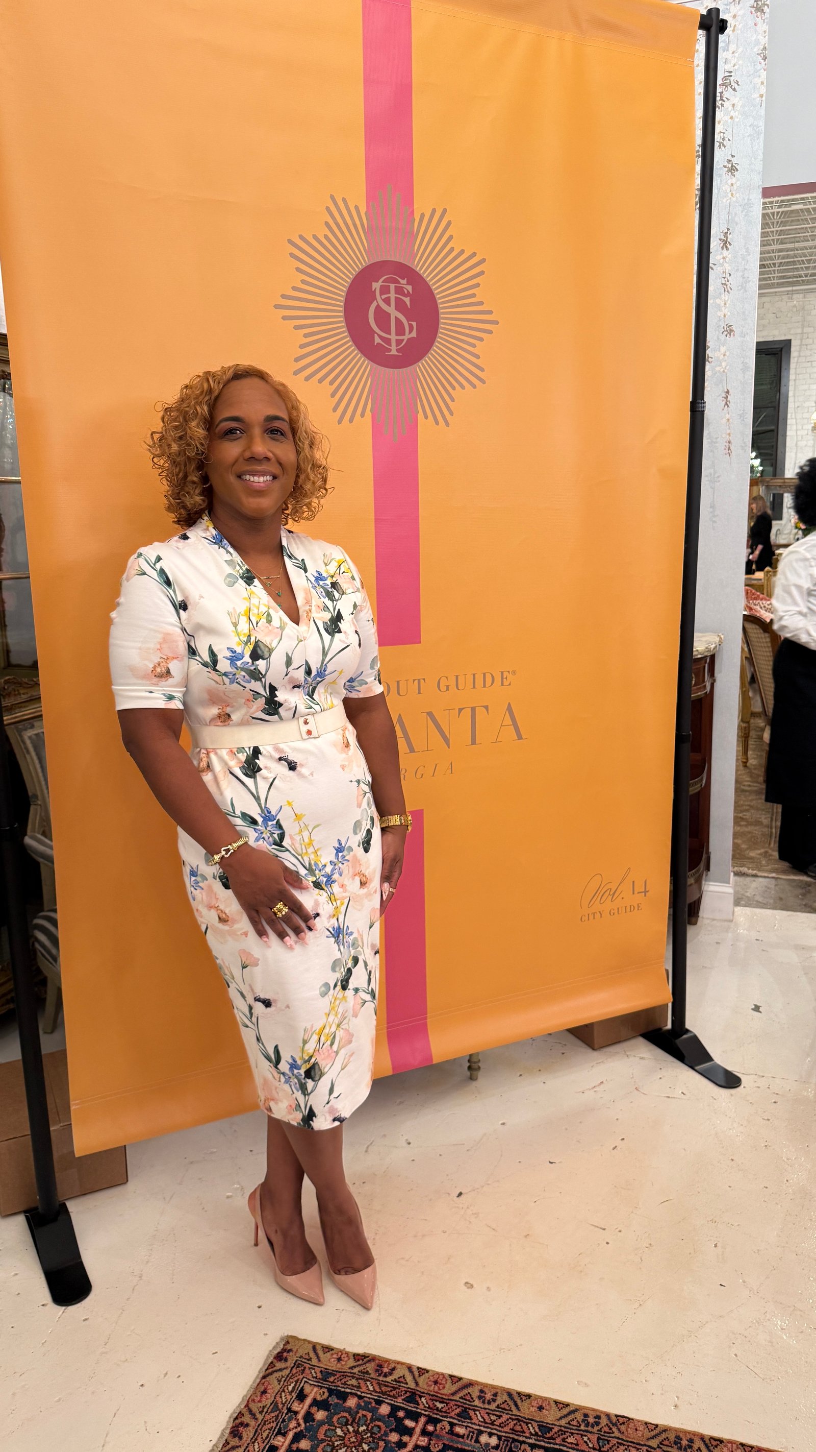A smiling woman in a floral dress poses in front of an orange Atlanta city guide banner inside an antiques showroom.