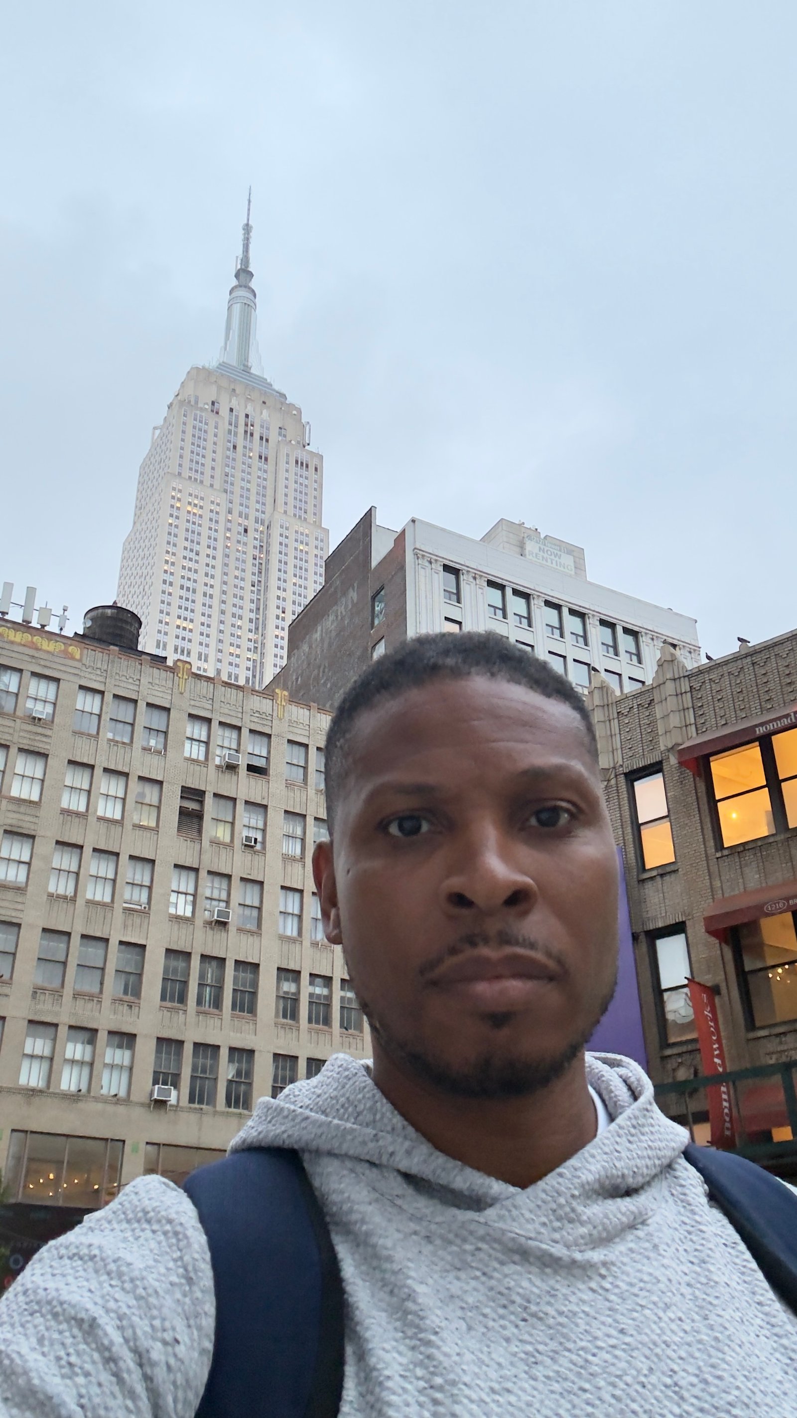 A man in a grey knit hoodie with a navy backpack takes a selfie on a Manhattan street, with the Empire State Building rising behind him against an overcast sky.