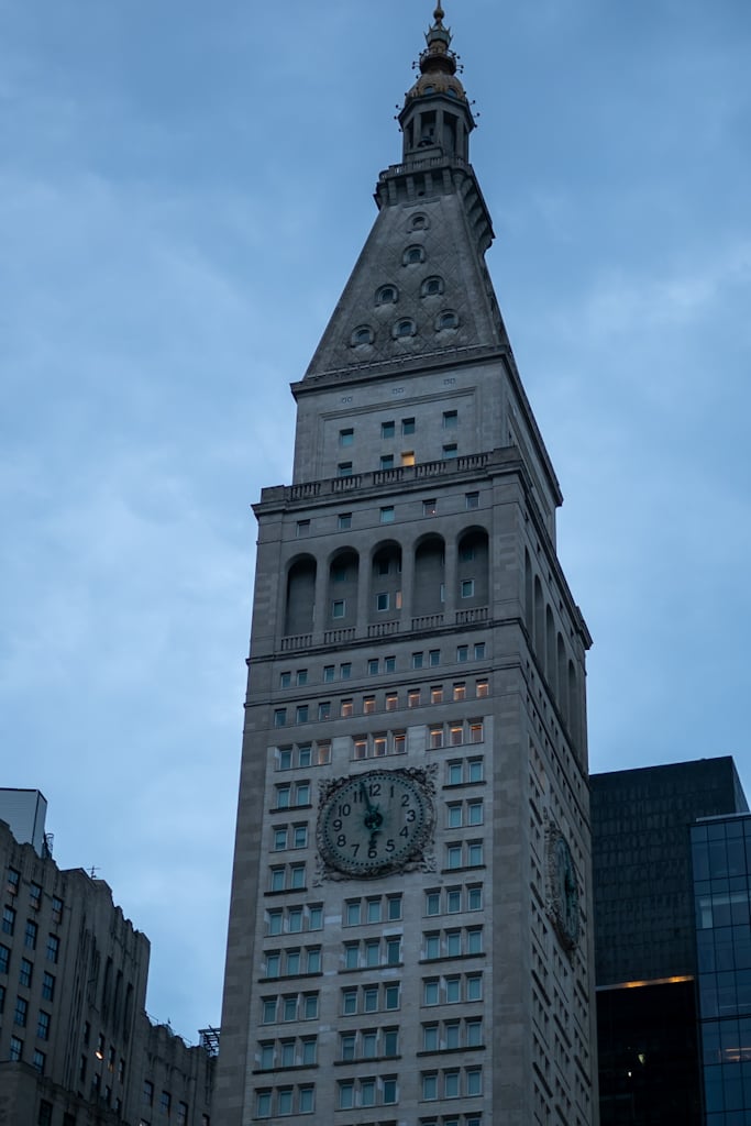 Tall grey stone clock tower with a pointed roof, round windows, a gilded finial and a large clock face, rising against a blue early-morning sky.