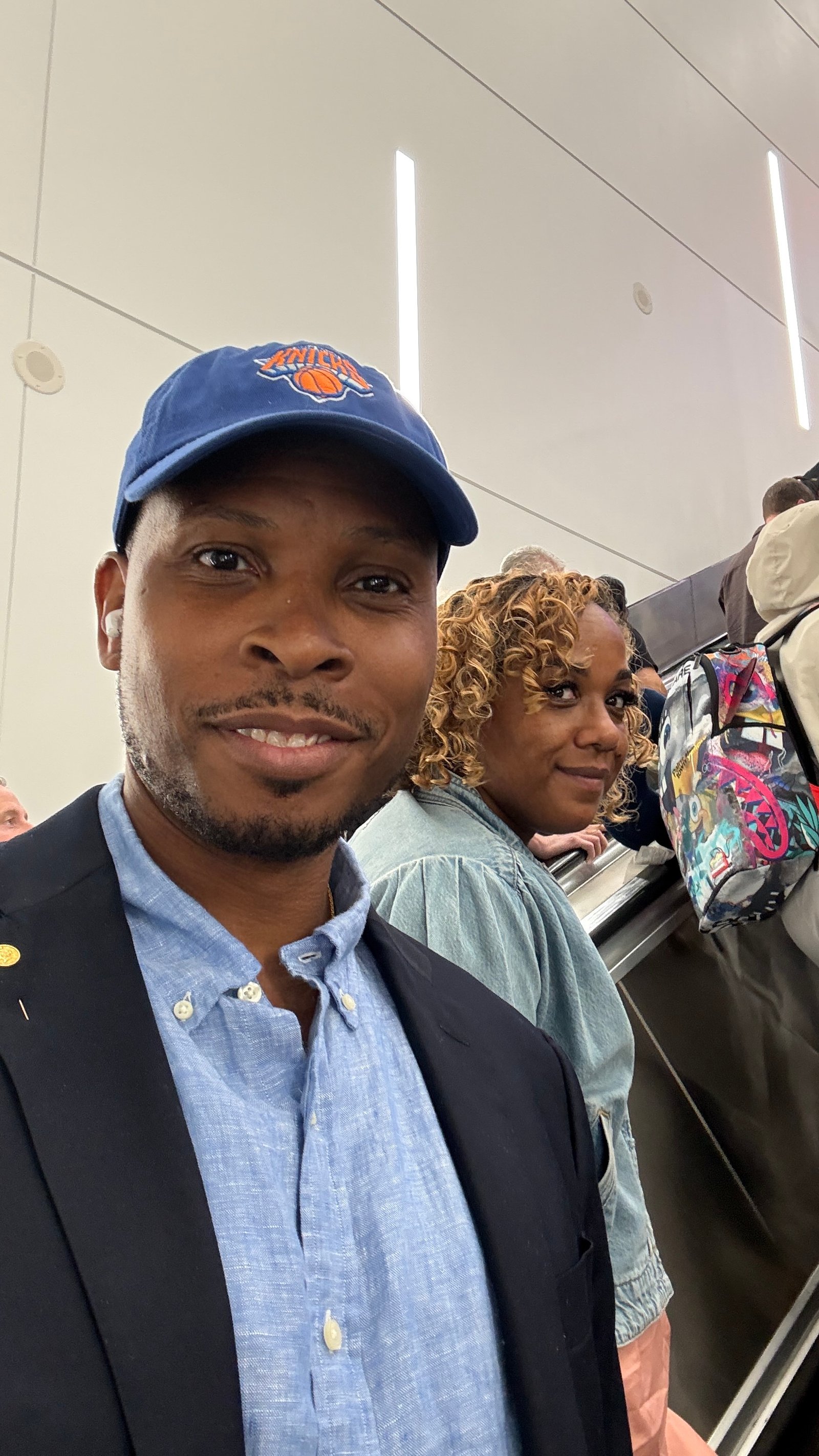 A man in a navy Knicks cap, light blue shirt and dark blazer takes a selfie on an escalator, with a woman with curly honey-blonde hair in a denim top behind him.