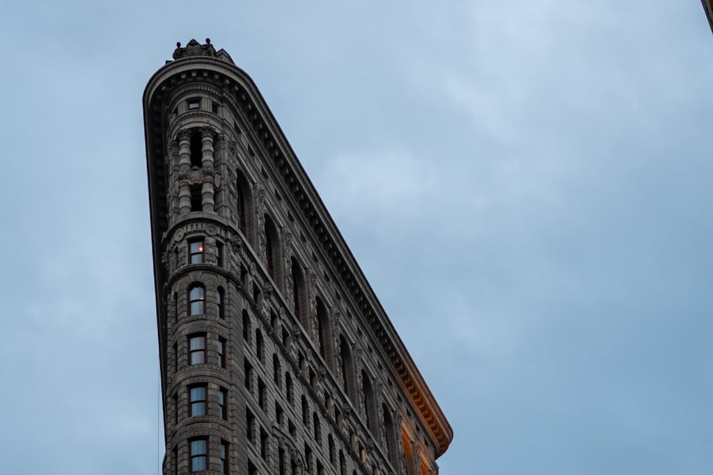 Close low-angle view of the narrow stone prow of the Flatiron Building, with carved ornament, arched windows and a small finial against a blue-grey sky.