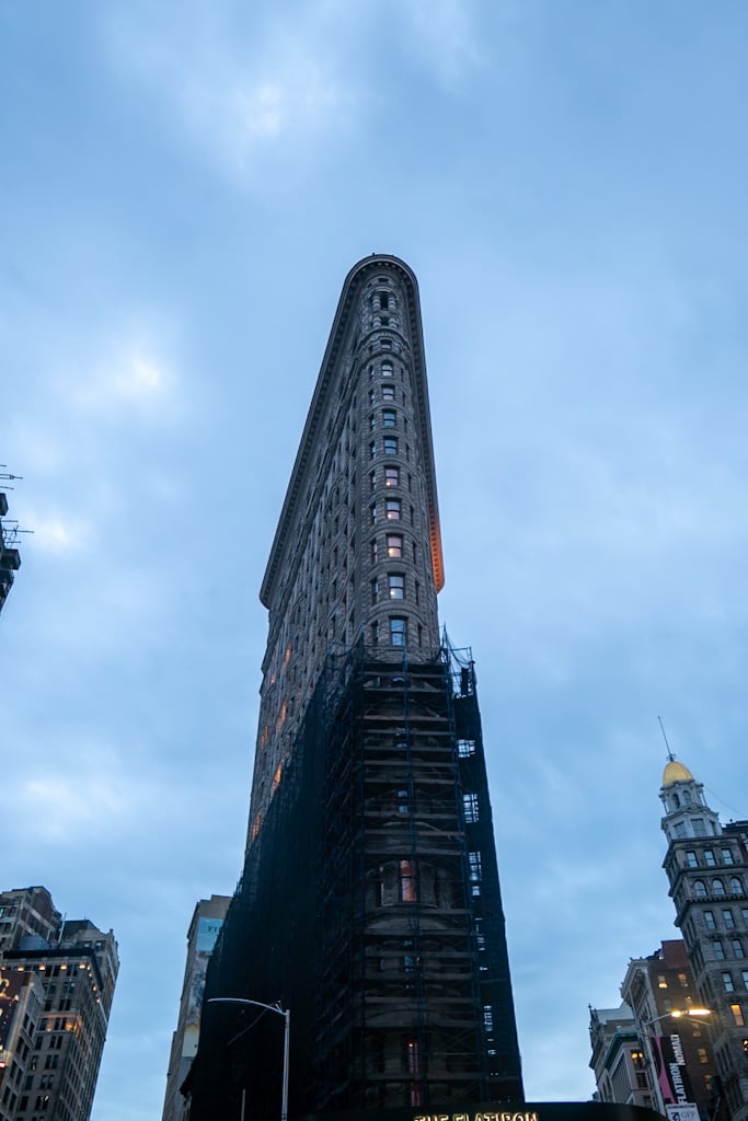 The Flatiron Building seen from street level in early blue light, its lower half wrapped in black scaffolding and netting, with warm light at the top edge.