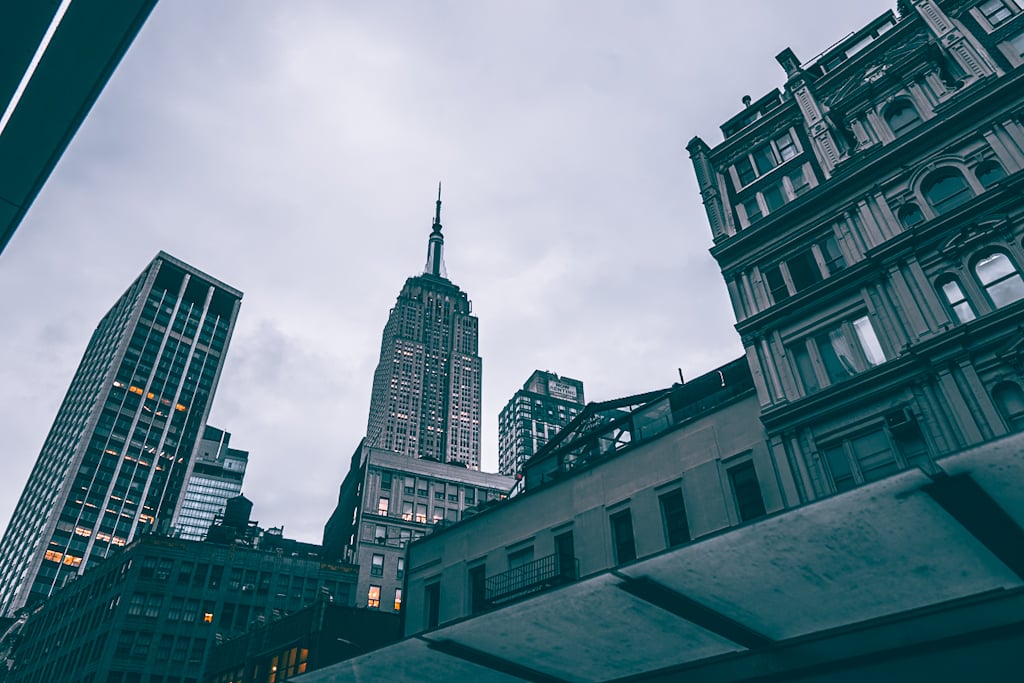 Low-angle view of the Empire State Building's spire on an overcast morning, framed by glass towers on the left and an ornate stone building with arched windows on the right.