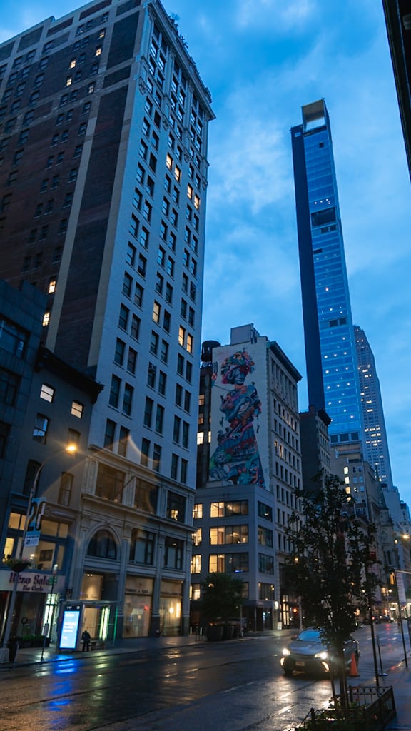 Wet city street in early blue light, with a tall stone office building, a colorful mural on a mid-rise facade, a slim glass tower, and a car with its headlights on.