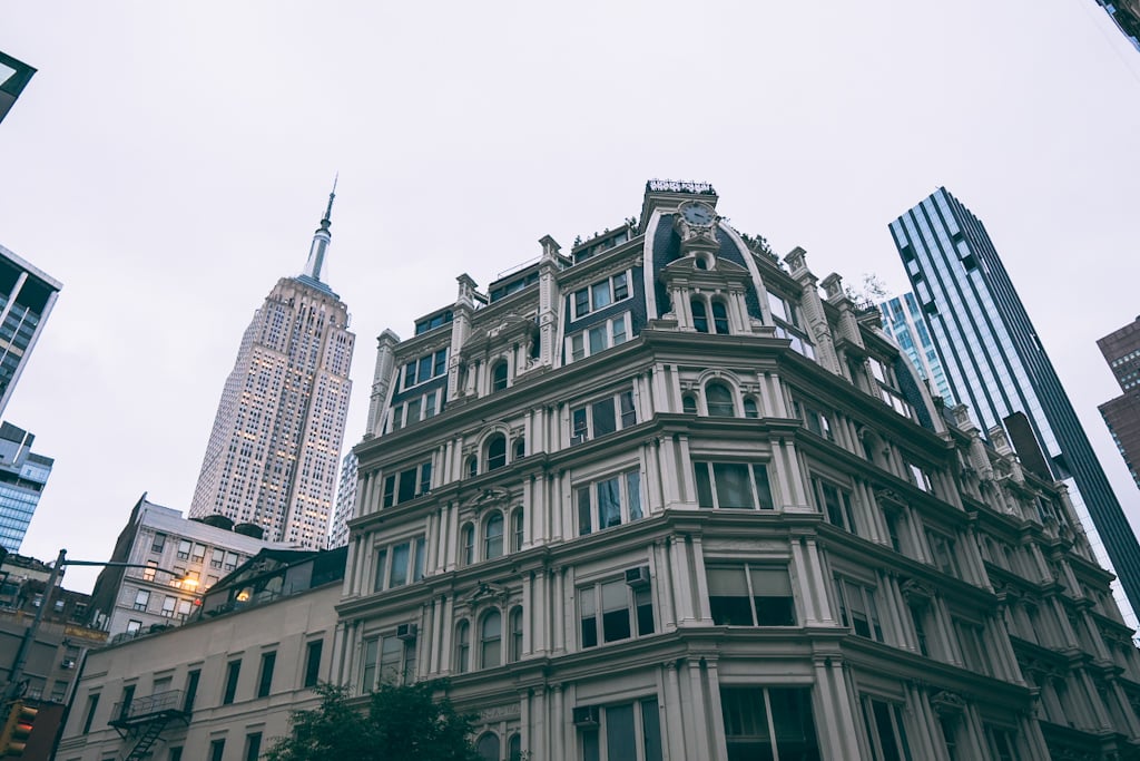 Ornate cream stone building with arched windows and a small clock in its corner tower, with the Empire State Building rising behind it under a grey sky.