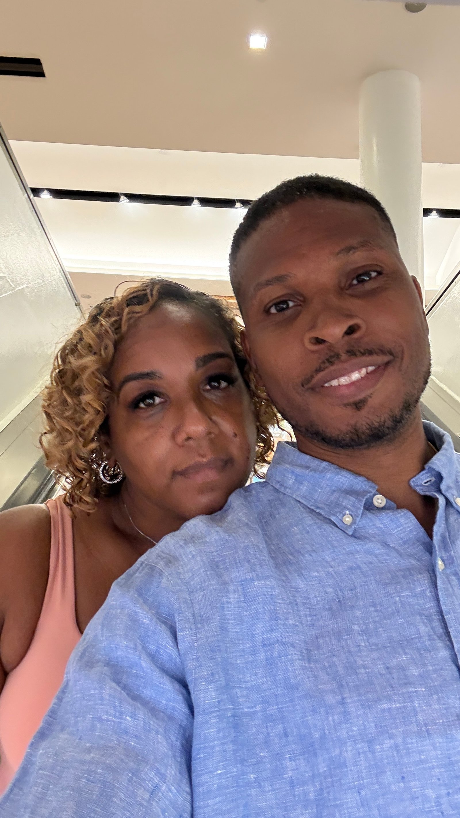 A man in a light blue linen shirt and a woman with curly honey-blonde hair and hoop earrings lean their heads together for a selfie on an escalator in a bright white interior.