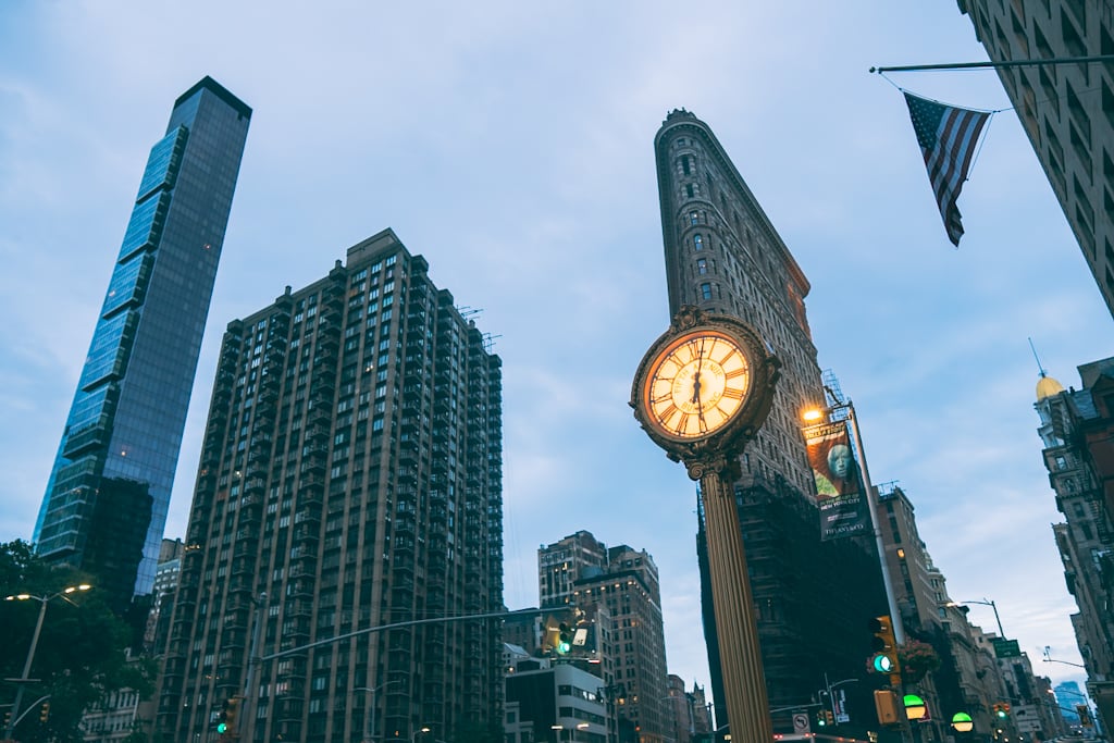 Lit ornate street clock on a tall pillar in front of the Flatiron Building, with modern towers to the left and an American flag hanging at upper right.