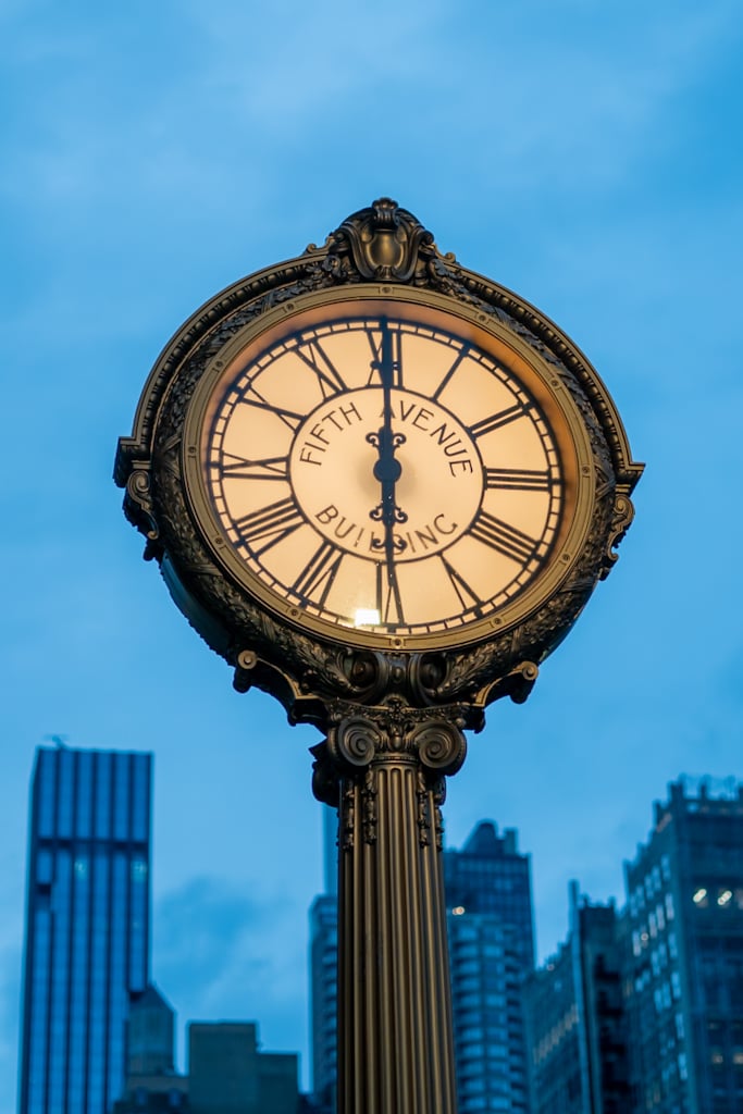 Close vertical view of a bronze pedestal street clock with Roman numerals and the words Fifth Avenue Building on its lit face, against a blue sky with towers behind.