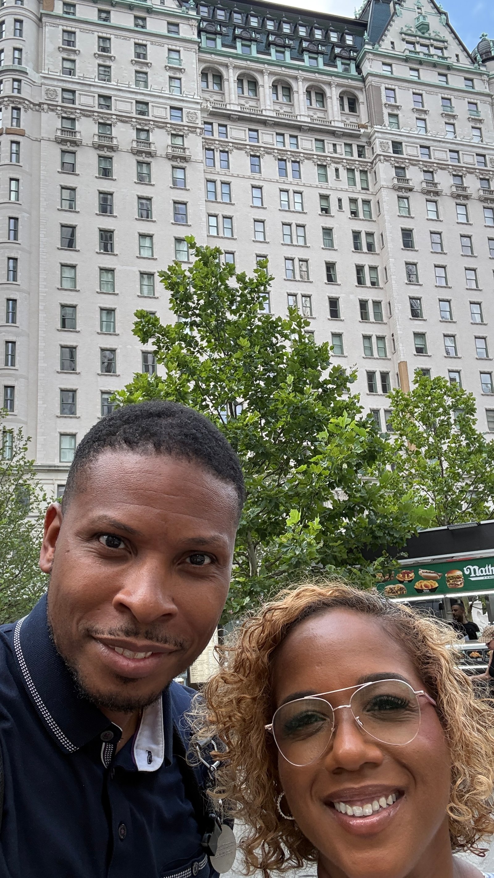 A man in a navy polo and a woman with curly honey-blonde hair and large glasses take a selfie on a city sidewalk in front of a tall cream stone building with green copper trim, trees and a hot dog stand sign at the edge of the frame.